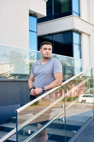 Gri tişörtlü, siyah şortlu, beyaz tenli yakışıklı, esmer adam iş merkezinin arka planında duruyor.