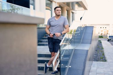 Gri tişörtlü, siyah şortlu, beyaz tenli yakışıklı, esmer adam iş merkezinin arka planında duruyor.