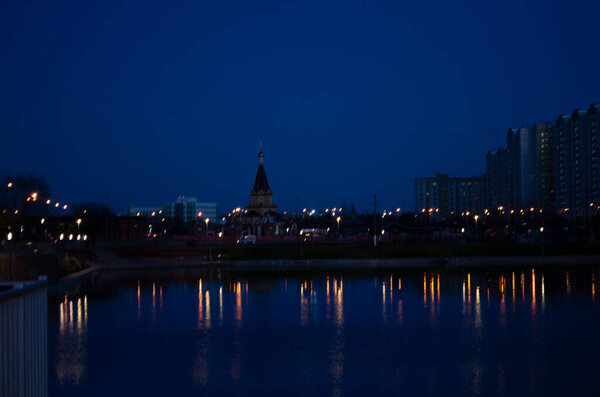 Night city landscape, lake in the foreground, church and buildings with night lights in the background. Display in water