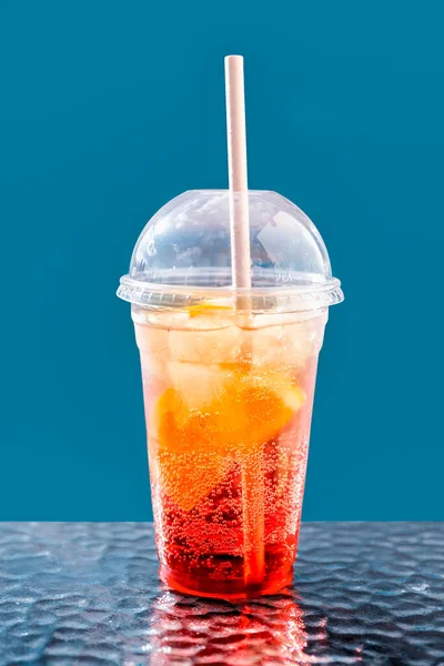 Fruitcocktail met ijs in een plastic glas op een blauwe achtergrond. Zomer drankje. Niet-alcoholische sinaasappel en aardbeiencocktail. Concept meeneem. — Stockfoto