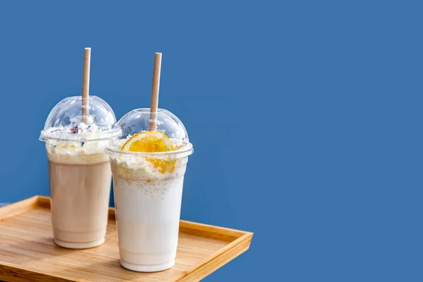 Frappe ve tek kullanımlık plastik bardakta milkshake, mavi arka planda kapaklı. Yaz içkileri. Götürün..