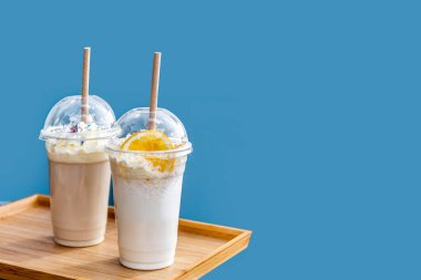 Frappe ve tek kullanımlık plastik bardakta milkshake, mavi arka planda kapaklı. Yaz içkileri. Götürün..