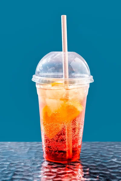 Cóctel de frutas con hielo en un vaso de plástico sobre un fondo azul. Bebida de verano. Cóctel de naranja y fresa sin alcohol. Concepto para llevar. Imagen De Stock