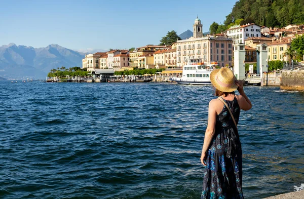 Una mujer en un sombrero de paja está de pie con su espalda y mira el lago Como lago y Bellagio. Ciudad en la ladera de las montañas. Hermoso paisaje marino de verano con fondo borroso. Tarjeta. Cartel. Imagen De Stock
