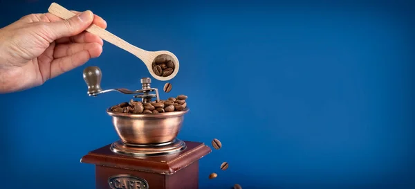 Uma mão polvilhe grãos de café em um moedor de café manual com uma colher. Fundo azul escuro. — Fotografia de Stock