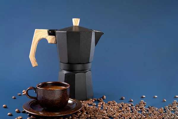 Geiser preto máquina de café, grãos de café e uma xícara com café em um fundo azul. — Fotografia de Stock