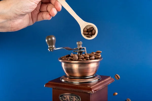 Uma mão polvilhe grãos de café em um moedor de café manual com uma colher. Fundo azul escuro. — Fotografia de Stock