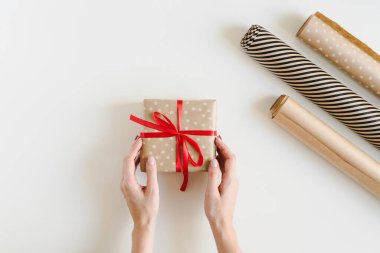 Bayanın elleri, kraft polka kağıdına sarılmış kırmızı kurdeleli ve beyaz masadaki kağıt rulolara sarılı Noel hediyesi kutusunu tutuyor. Çevre dostu Noel hediyesi paketleme fikri