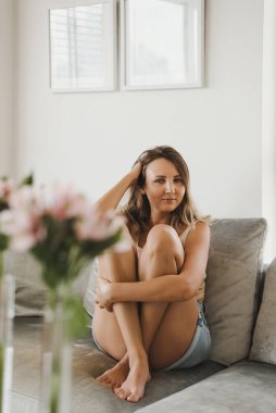 Dairesinde gri bir kanepede oturan genç bir kadın. Ev kavramında rahatlamak. Rahatlık ve rahatlık. Evde kal.