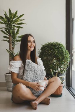 Mutlu ve gülümseyen genç bir kadın, elinde bir fincan kahveyle kapalı bahçenin yanında oturmuş pencereden dışarı bakıyor. Evde rahatla, pozitif tavır ya da kişisel bakım kavramı