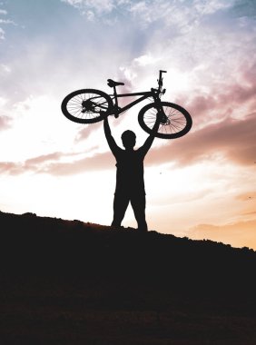 Dağ bisikletçisinin silueti başarılı olduğu için motoru mutlu bir şekilde kaldırır. Akşamları yüksek dağlardaki yarışmalardan, rengarenk..