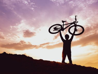 Dağ bisikletçisinin silueti başarılı olduğu için motoru mutlu bir şekilde kaldırır. Akşamları yüksek dağlardaki yarışmalardan, rengarenk..