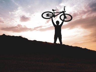 Dağ bisikletçisinin silueti başarılı olduğu için motoru mutlu bir şekilde kaldırır. Akşamları yüksek dağlardaki yarışmalardan, rengarenk..