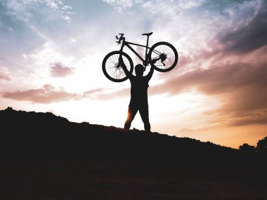 Dağ bisikletçisinin silueti başarılı olduğu için motoru mutlu bir şekilde kaldırır. Akşamları yüksek dağlardaki yarışmalardan, rengarenk..