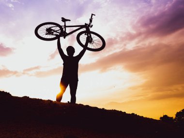 Dağ bisikletçisinin silueti başarılı olduğu için motoru mutlu bir şekilde kaldırır. Akşamları yüksek dağlardaki yarışmalardan, rengarenk..