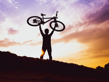 Dağ bisikletçisinin silueti başarılı olduğu için motoru mutlu bir şekilde kaldırır. Akşamları yüksek dağlardaki yarışmalardan, rengarenk..