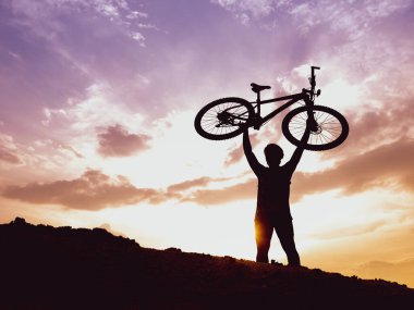 Dağ bisikletçisinin silueti başarılı olduğu için motoru mutlu bir şekilde kaldırır. Akşamları yüksek dağlardaki yarışmalardan, rengarenk..