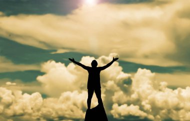 Kayaların üzerinde duran silüet bir adamın elleri havada ve arka planda bulutlu bir gökyüzü