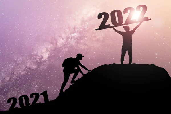 欢迎快乐的圣诞和快乐的新年在2022年 轮廓人从2021悬崖到2022悬崖与云天和阳光 男子举着2022年的旗帜在山顶上 祝2022年新年快乐 — 图库照片