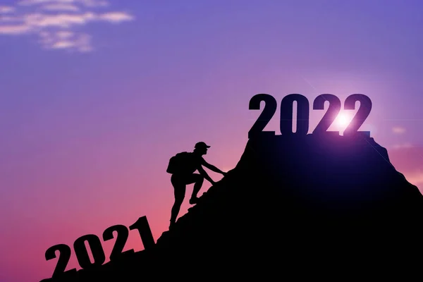 Welcome merry Christmas and happy new year in 2022,Silhouette Man from 2021cliff to 2022 cliff with cloud sky and sunlight. Man holding 2022 banner on top of mountain. Happy New Year 2022 concept. Rock climbing man finds 2022 sign on top of mountain.