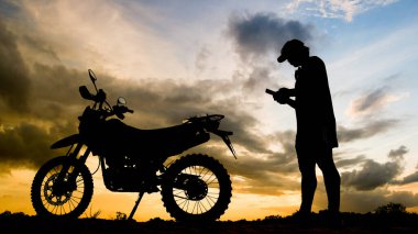 The silhouette of an adventurous traveler man looking at mobile phone with motocross motorcycle in the evening