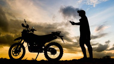 The silhouette of an adventurous traveler man looking at mobile phone with motocross motorcycle in the evening