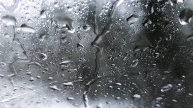 Cam bir pencerenin üzerindeki yağmur damlalarının yakın plan görüntüsü bulanık ve dengesiz bir arkaplan karşısında soyut desenler oluşturuyor..