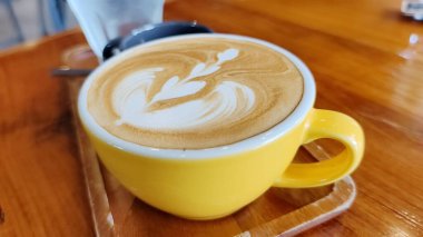 Canlı sarı bir fincanda, ahşap bir masa üzerinde kalp ve yaprak tasarımı sergilenen, çok güzel hazırlanmış bir latte sanatının yakın çekimi..