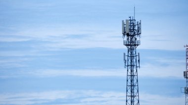 Bir iletişim kulesi mavi gökyüzüne karşı ayakta duruyor. Dağılmış bulutlar modern teknolojiyi ve bağlantıyı simgeliyor..