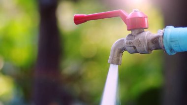 Açık deniz musluğu, kırmızı saplı, akan su, ve güneş ışığında yeşil bahçe arka planı.