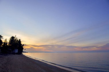 Gün batımında huzurlu bir plaj sahnesi. Renkli gökyüzü ve sakin okyanus dalgaları. Seyahat ve rahatlama temaları için mükemmel..