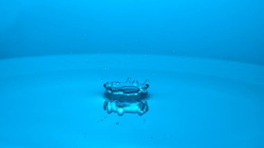 Su damlacığının büyüleyici bir görüntüsü dingin mavi bir yüzeyde dalgalanmalar yaratarak sıvı dinamiğinin güzelliğini gözler önüne seriyor..