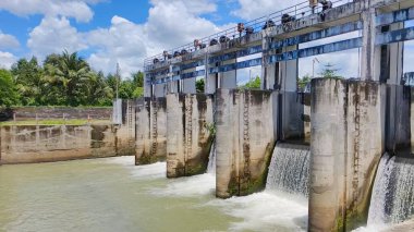 Su akan modern bir baraj, canlı mavi bir gökyüzü ve yemyeşil üzerine kurulmuş..