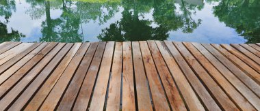 Sakin bir gölün kenarındaki ahşap rıhtımın huzurlu manzarası yeşil ağaçları ve mavi gökyüzünü yansıtıyor, doğa ve rahatlama temaları için mükemmel..