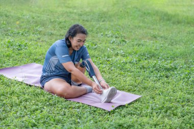 Spor kıyafetli, protez bacaklı Asyalı bir kadın yoga minderinde oturuyor ve parkta ayakkabı bağı bağlıyor.