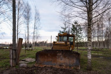 Sürücüsüz bir çiftlikteki açık havada ağaçların arasında paslı sarı bir traktör. Tarım makineleri