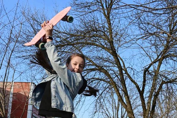 デニムとピンクのスニーカーの美しい陽気な女の子がジャンプし 彼女の頭の上にロングボード ペニーボードを保持しています 国際スケートボードの日 スポーツと開発の概念 ライフスタイル — ストック写真