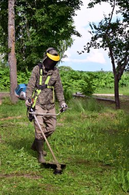 Elinde çim biçme makinesiyle özel korumalı giysiler içinde çalışan işçiler çim biçiyor. Bahçıvan çimleri biçme makinesi, çim biçme makinesi veya çim biçme aracı ile keser