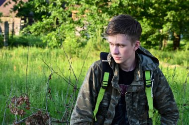 Koruyucu tulumlu genç yorgun işçi dinleniyor, sıkı çalışmadan sonra arka planda yeşil ağaçların karşısında oturuyor. Çalışma ve dinlenme zamanı.