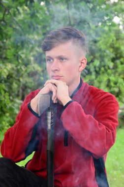 Ormanda oturan, ellerini baltaya yaslayan ciddi bir gencin portresi. Ateşten çıkan dumanın etrafında. Doğaya giden yol. Macera kavramı