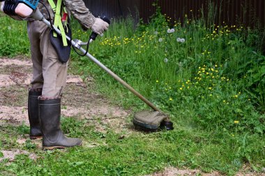 Elinde çim biçme makinesi olan özel korumalı elbiseli bir adam çim biçiyor. Bahçıvan çimenleri budayıcı, çim biçme makinesi ya da güneşli bir günde çim biçmek için elektrikli bir araç ile biçer.