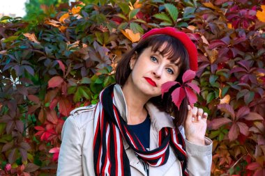 Kırmızı bereli zarif esmer ve dudaklarında parlak kırmızı rujla elinde bir sonbahar yaprağı tutuyor. Yakın plan. Merhaba sonbahar! Açık hava eğlencesi