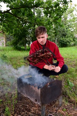 Genç adam et pişiriyor, mangalda mangal yapıyor. Dışarıda bekliyor. Barbekü yeşil çimlerin üzerinde yer alır. Arka planda ağaçlar var. Boş zaman geçirme kavramı, ortak barbekü, yaz mevsimi