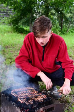 Genç adam et pişiriyor, doğada kömürde barbekü yapıyor. Barbekü yeşil çimlerin üzerinde yer alır. Arka planda ağaçlar var. Boş zaman, yaz tatili ve barbekü kavramı