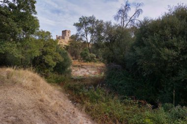 Şato kalıntıları yeşillik ve çalılarla çevrili arka planda yükselirken, huzurlu bir dere, dağınık bulutlarla dolu yumuşak bir akşam gökyüzünün altında akıyor..