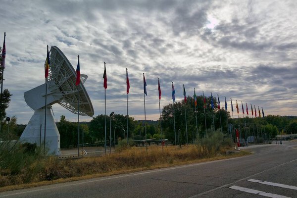 Значительно стоит большая спутниковая тарелка, окруженная флагами разных стран, выстилающими тропу. Облачное небо добавляет драматический фон к сцене, улучшая мирную обстановку.