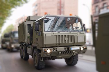 Şehir caddesi boyunca bir konvoyda ağır askeri kamyonlar hareket ederken görülüyor. Araçlar yeşil, şehir manzarası ile harmanlanmış ve askeri varlıklarını gösteriyor..