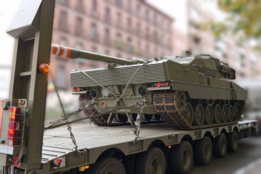 Bir askeri tank, zırhlı vücudunu ve uzun namlulusunu gösteren bir kamyonete güvenli bir şekilde yükleniyor. Sahne, arka plandaki kentsel binaları yakalıyor ve yoğun bir şehir yaşamına dair ipuçları veriyor..