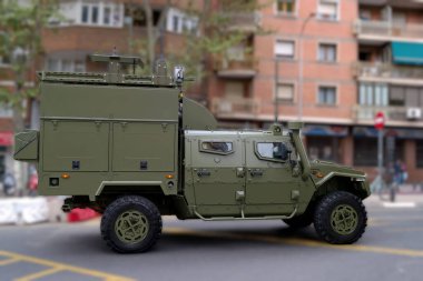 Gelişmiş teknolojiyle donatılmış bir askeri araç bir şehir caddesinde konuşlandırıldı. Çevresindeki binalar ve ağaçlar tipik bir şehir atmosferi yaratıyor..