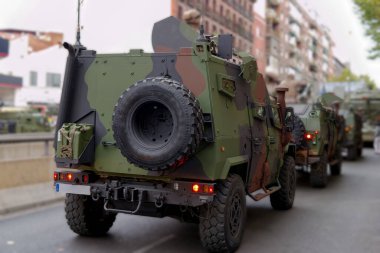 Birkaç askeri araç, kamuflaj desenlerini sergilemek için bir şehir caddesine park etmiş durumda. Güneş, araçların mevcudiyetini vurgulayarak kentsel arkaplanı vurguluyor.